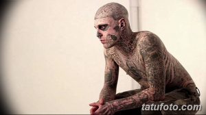Фото тату: мужчина с полностью татуированным лицом и телом в стиле биомеханика, сидящий в задумчивой позе.